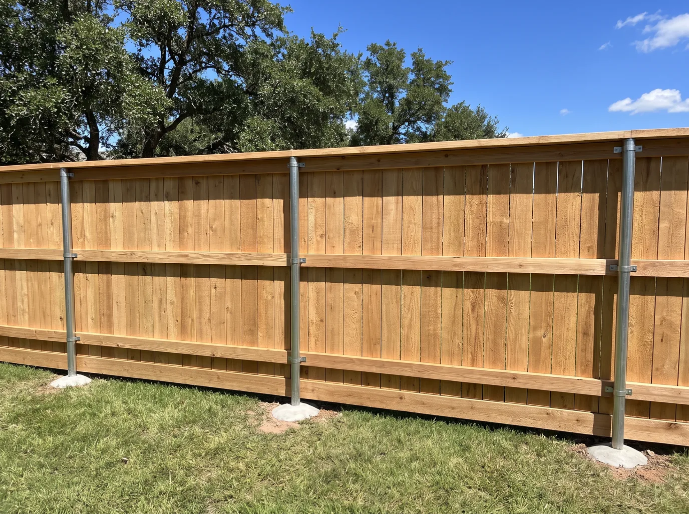 The inside face of a new cedar privacy fence: galvanized steel posts in concrete footings, cedar rails held on with brackets, a kickboard, and a cap along the top